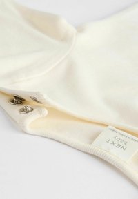Cremfarbener Baby-Body aus weichem Stoff mit Druckknöpfen am Boden und einem Etikett mit der Aufschrift "NEXT Baby."