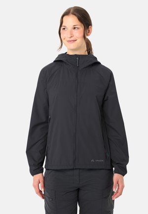 Donna che indossa una giacca softshell nera con cappuccio, cerniera frontale e tasche laterali, in piedi davanti a uno sfondo chiaro uniforme.