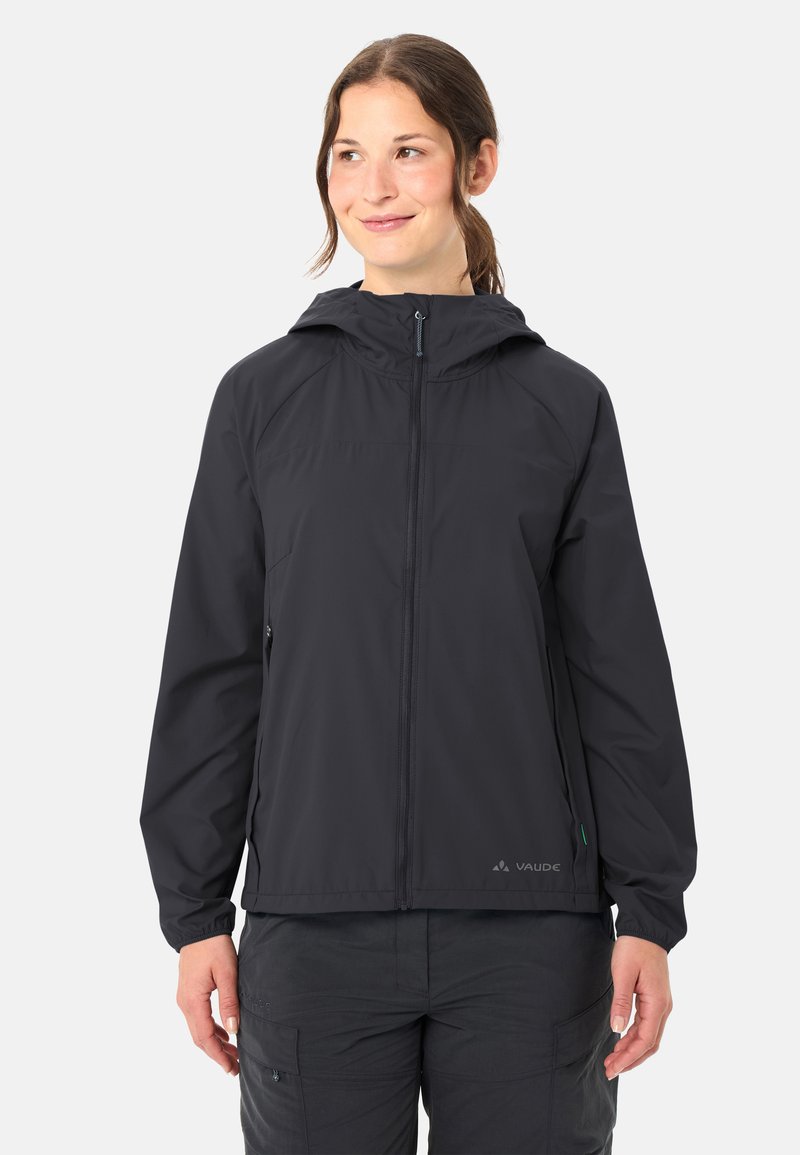 Donna che indossa una giacca softshell nera con cappuccio, cerniera frontale e tasche laterali, in piedi davanti a uno sfondo chiaro uniforme.