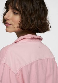 Lichtroze shirt met een rafelende kraag, zichtbare stikseldetails en een losse pasvorm. De stof lijkt zacht met een lichte textuur.