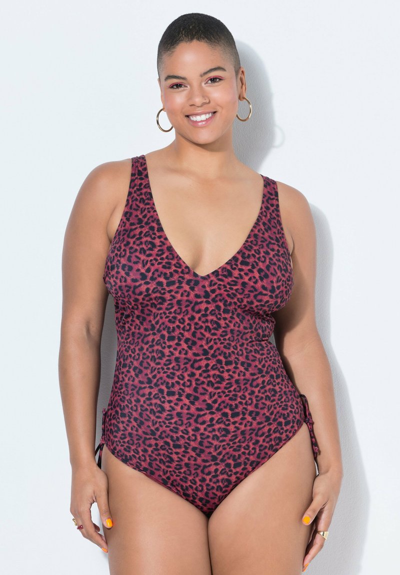 Uśmiechnięta osoba nosząca duże kolczyki-koła i czerwony jednoczęściowy strój kąpielowy w cętki lamparta na jednolitym, jasnym tle.