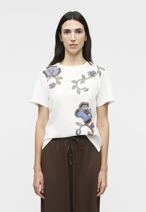 Femme aux longs cheveux foncés portant une chemise blanche à manches courtes avec des motifs floraux bleus et bruns et un pantalon marron à cordon de serrage.