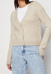 Beige wool cardigan with a V-neck and long sleeves, featuring four decorative buttons and ribbed cuffs. Worn over a white t-shirt and blue jeans.