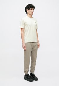 Gaiši bēša īsroku t-krekls ar nelielu logotipu, kopā ar gaiši pelēkām joggerstāvas biksēm un melnām sporta kurpēm, ar plūstošu dizainu.