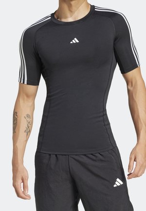 Mann trägt schwarze, eng anliegende, kurzärmelige Sportshirt mit weißen Streifen an den Ärmeln und passende schwarze Shorts mit Logo am linken Bein.
