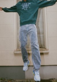 Zelená mikina s potiskem, světle modré manšestrové džíny a bílé tenisky s modrými a zelenými akcenty; osoba ve skoku před zdí.