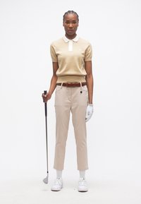 Γυναίκα με μπεζ πόλο πουκάμισο και παντελόνι κρατώντας ένα γκολφ μπαστούνι, φορώντας λευκό γάντι γκολφ και λευκά αθλητικά παπούτσια, στέκεται σε λευκό φόντο.