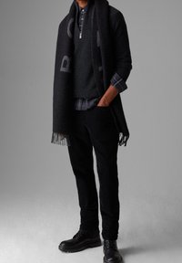 Écharpe noire à franges, pull gris, chemise à carreaux et pantalon en velours côtelé noir, associés à des bottes noires à lacets. Look simple et superposé.