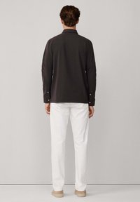 Maglietta a maniche lunghe di colore grigio scuro con colletto, polsini abbottonati, abbinata a pantaloni bianchi e scarpe beige, vista da dietro.
