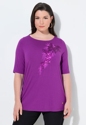 Kvinde med mørkt hår iført lilla, kortærmet top med diagonalt blomster-sequin-design, kombineret med sorte bukser på ensfarvet hvid baggrund.