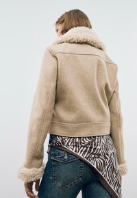 Beige zkrácená bunda s chlupatým bílým límcem a manžetami, doplněná tmavými džínami a hnědým tričkem se zebřím vzorem pod ní.