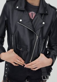 Chaqueta de moto de imitación cuero negra con cremalleras plateadas, botones a presión y un cinturón. Presenta bolsillos laterales y en el pecho, solapas anguladas y un acabado texturizado.