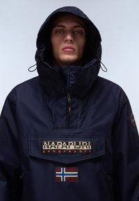 Rompevientos marino con capucha, que presenta un bolsillo frontal y el logotipo "NAPAPIJRI". Incluye un parche con un diseño de bandera. Textura suave.