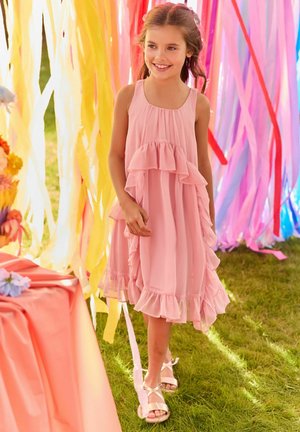 Junges Mädchen in einem rosa, gerafften Kleid lächelt und läuft auf Gras mit bunten hängenden Girlanden im Hintergrund.