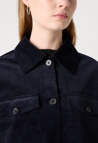 Veste chemise en velours côtelé bleu marine avec un col pointu, deux poches avant et des boutons noirs. Texture lisse avec un tissu à côtes.