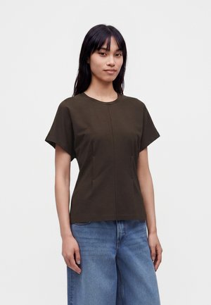 Frau mit langen dunklen Haaren, die ein dunkelbraunes T-Shirt mit kurzen Ärmeln und hellblaue, weite Jeans trägt, steht vor einem schlichten Hintergrund.