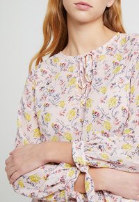 Blusa floreale di un delicato rosa pallido, con fiori gialli e rossi, vestibilità ampia, scollo rotondo con dettaglio a fiocco e polsini delle maniche arricciati.