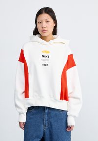 Haut à capuche blanc avec des accents rouges, arborant un imprimé logo sur le devant et le texte 1972. Confectionné dans un tissu doux avec une coupe décontractée et une poche kangourou.