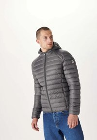 Grå puffjacka med huva, dragkedja och ribbat mönster. Har en logotyp på vänster arm. Bärs över blå jeans.