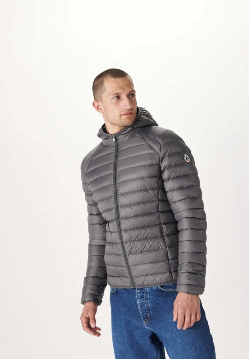 Grå puffjacka med huva, dragkedja och ribbat mönster. Har en logotyp på vänster arm. Bärs över blå jeans.
