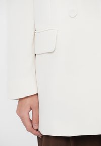 Biely blejzer s precíznym strihom, vybavený jedným gombíkom, našitou vreckovou záplatou na rukáve a hladkou, textúrovanou látkou.