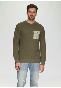 Oliwkowy sweter z prążkowanej dzianiny z długimi rękawami, ozdobiony jasnooliwkową kieszenią na lewej piersi z czarnym guzikiem. W połączeniu z jasnoniebieskimi dżinsami.
