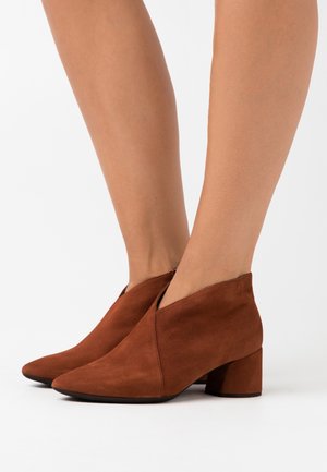 Botas de ante marrón hasta el tobillo con tacones cuadrados, llevadas sobre piernas desnudas contra un fondo blanco.