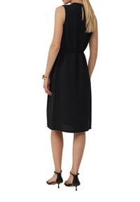 Robe noire sans manches avec une taille cintrée, une silhouette arrivée au genou et un détail en forme de trou de serrure au dos. Le tissu a une apparence texturée.