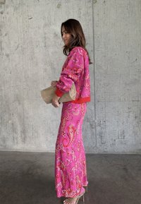 Γυναίκα με ροζ ρούχο με πασλέ σχέδιο κρατώντας μπεζ τσάντα φάκελο, όρθια πλάγια σε τοίχο με υφή από σκυρόδεμα, φοράει πέδιλα με λουράκια.