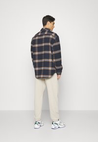 Camicia di flanella a quadri blu navy con righe beige e azzurre chiare, maniche lunghe e colletto button-down, abbinata a pantaloni beige chiaro e sneakers.