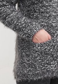 Grå texturerad tröja med en fluffig look, med långa ärmar och en ficka fram, som visar en blandning av ljusa och mörka grå fibrer.