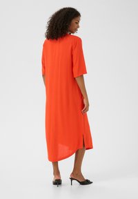 Robe orange à mi-longueur avec des manches courtes, des fentes latérales et une coupe droite. Portée avec des mules noires à talons et stylisée dans un cadre minimaliste.
