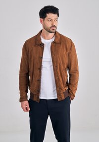 Man draagt een bruine suède jas over een wit t-shirt en donkere broek, staat met één hand in zijn zak en kijkt naar links.