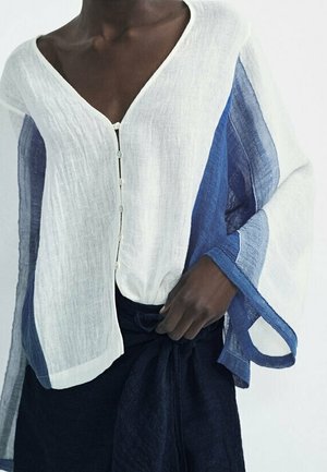 Personne à la peau foncée portant un chemisier blanc boutonné avec de larges manches à rayures bleues et une jupe noire nouée à la taille.