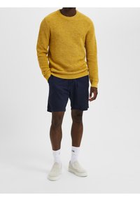 Gelber Strickpullover mit strukturiertem Muster, kombiniert mit marineblauen Shorts. Die Schuhe sind helle Slipper und weiße Knöchelsocken.