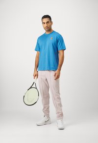 Blå bomull T-skjorte med en liten logo, kombinert med lyse rosa treningsbukser. Holder en tennisracket med et mønstret snorrist.