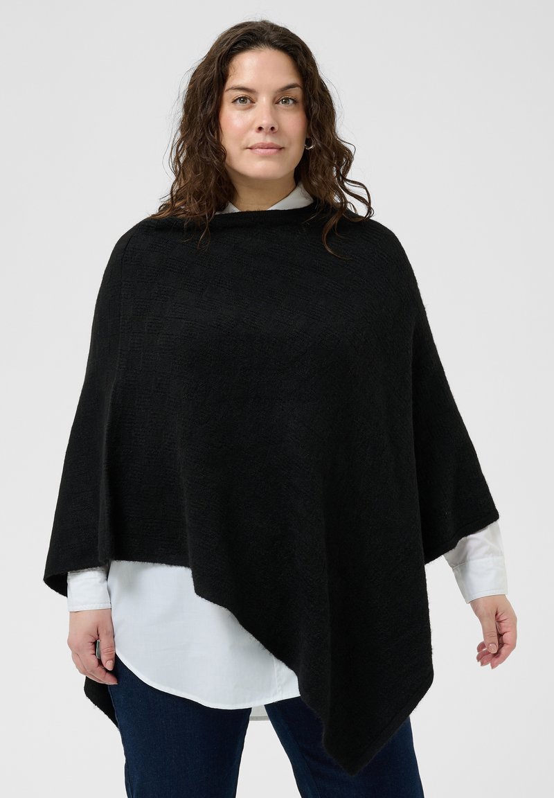 Femme aux cheveux bruns bouclés portant un poncho asymétrique noir sur une chemise blanche et un jean bleu, debout devant un fond uni.