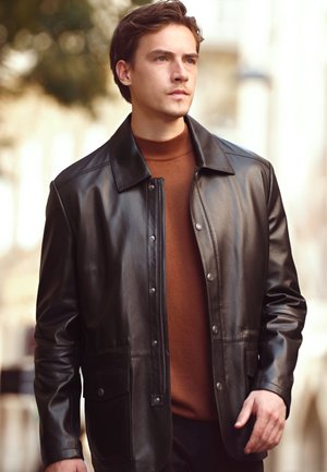 Jeune homme portant une veste en cuir noire sur un col roulé marron, debout en extérieur avec un arrière-plan urbain flou.