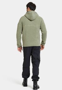 Grön fleecejacka med huva, strukturerad yta och svart detalj på övre delen av ryggen, bärs över svarta byxor och stövlar.