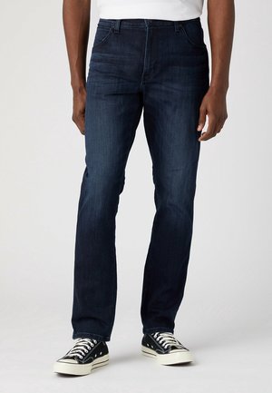 Man draagt donkerblauwe rechtdoor gesneden spijkerbroek en zwarte lage sneakers tegen een lichte achtergrond.