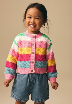 Lächendes junges Mädchen, das eine bunte gestreifte Strickjacke und Jeansshorts trägt und vor einem schlichten beigen Hintergrund steht.