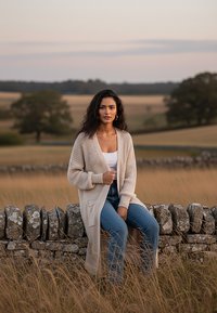 Długi, miękki, kremowy kardigan z teksturowanym wzorem dzianiny, z kieszeniami i luźnym krojem, noszony na białej bluzce i niebieskich dżinsach, siedzący na kamiennej ścianie.