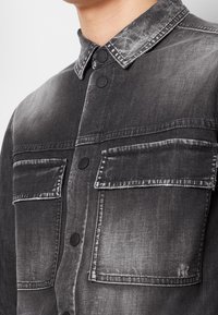 Camicia in denim nero con una texture sbiadita, collo con bottoni e due tasche sul petto con bottoni neri. Il tessuto ha un aspetto invecchiato.