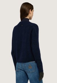 Námořnický modrý pletený svetr s texturou, vysokým límcem a štítkem s logem na zadní straně. Kombinováno s modrými džíny se vyšitým detailem.