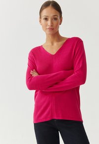 Rosa V-Ausschnitt-Pullover mit langen Ärmeln, gestrickter Textur und dezenten Nahtdetails, kombiniert mit dunklen Jeans. Weiches Material und lockere Passform.