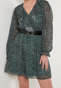 Vestido transparente verde y negro con manchas, de mangas largas, escote volante y un cinturón negro en la cintura, que presenta un diseño fluido.
