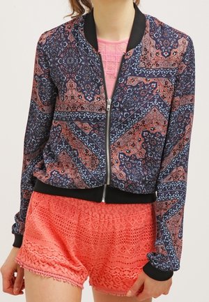 Femme portant une veste zippée avec motifs bleu marine et corail, avec bordure noire, et un short en dentelle corail vif.