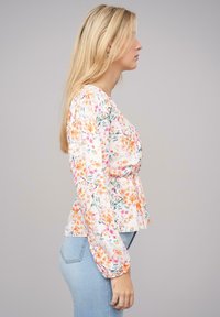 Blusa floreale in tessuto leggero con fiori arancioni e rosa, maniche lunghe a sbuffo e vita aderente, abbinata a jeans blu.