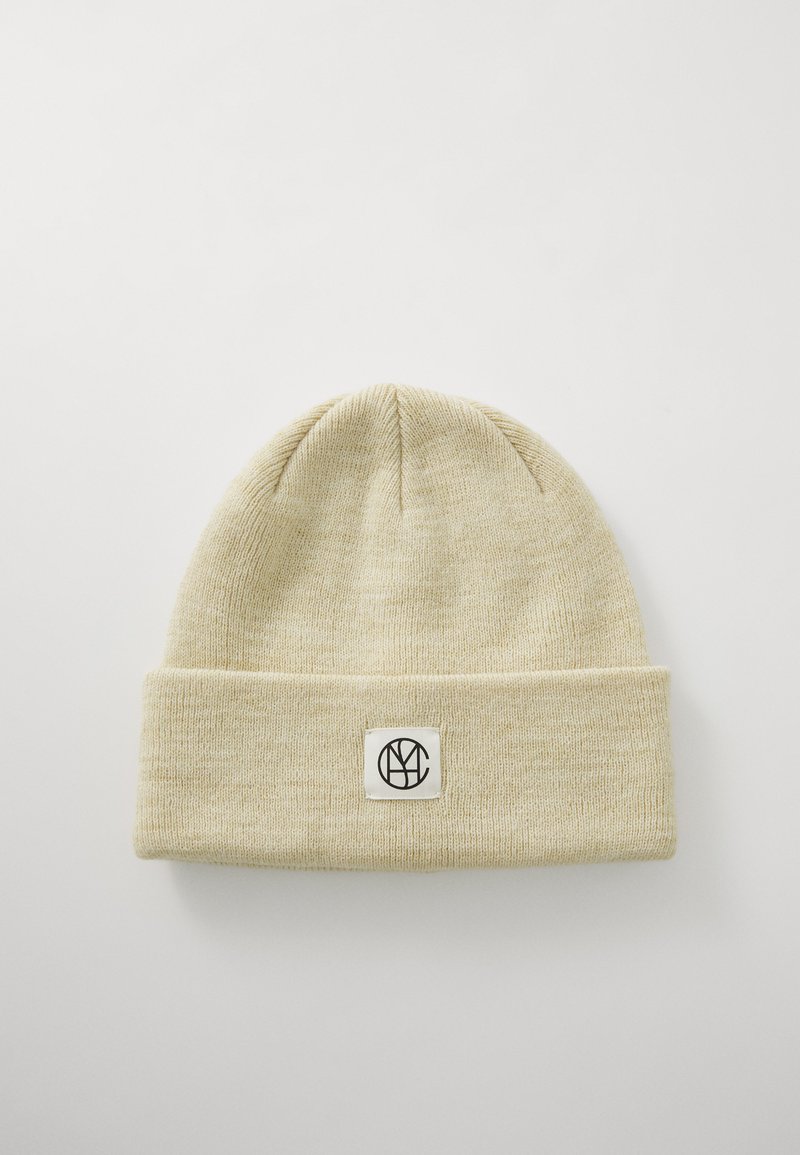 Lys beige strikket lue med brett, med en firkantet lapp med en svart logo i midten. Myk tekstur, enkelt design.