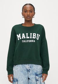Grüner Pullover mit dem weißen Schriftzug "MALIBU CALIFORNIA". Verfügt über einen Rundhalsausschnitt und eine lockere Passform, kombiniert mit hellblauen Denim-Cargohosen.